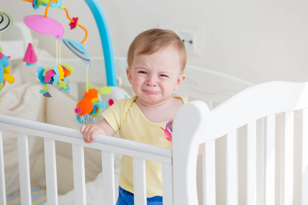 Portriat of 10 months old baby boy standing in cot and crying - Φωτογραφία, εικόνα