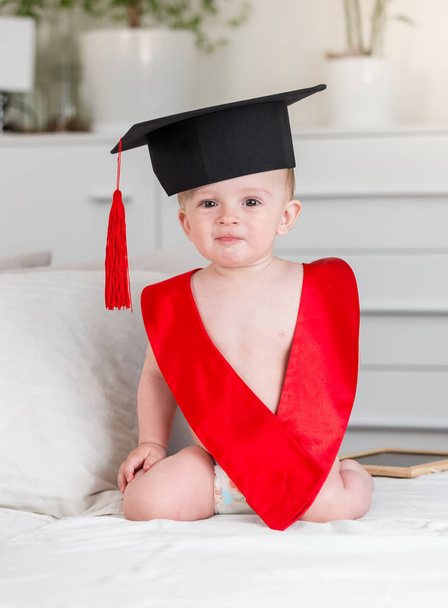 Retrato de niño adorable en sombrero de mortero y cinta roja mirando en cámara. Concepto de educación infantil temprana
 - Foto, Imagen