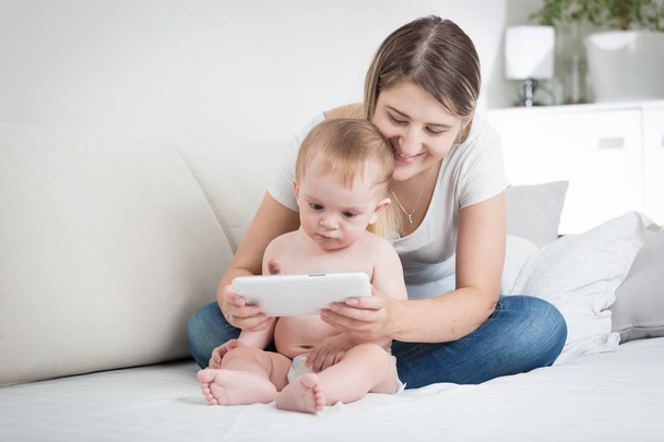 Young mother sitting on bed with her baby son and showing him cartoons on digital tablet - Foto, Bild