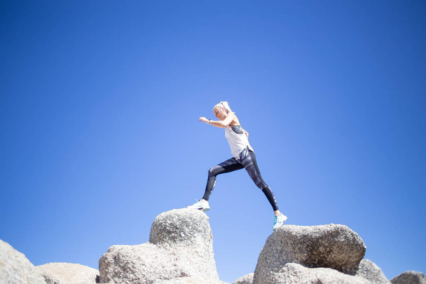 Beautiful blond model in sportswear jumping from rock to rock against bright blue sky at daytime - Photo, Image