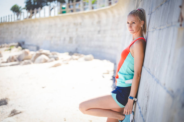 Beautiful fitness model in sportswear resting after workout on beach at daytime, brick wall background - Fotó, kép