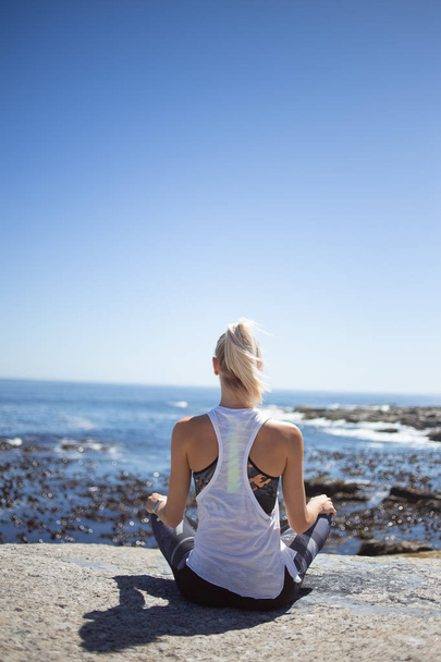 Blond female model meditating and doing yoga on granite rock overlooking ocean at daytime - 写真・画像