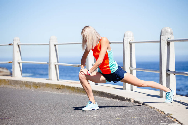 Young female fitness athlete stretching before outdoor running at daytime, ocean under blue sky background - Photo, Image