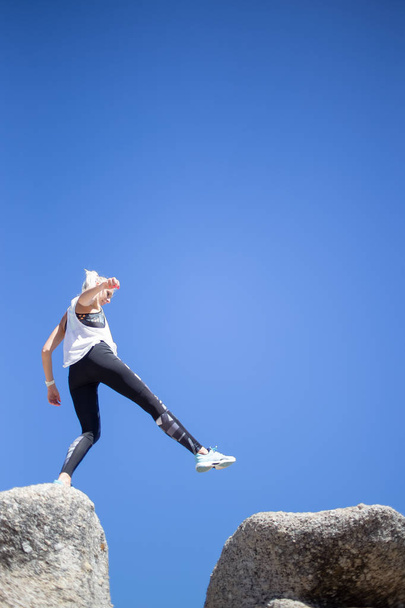 Beautiful blond model in sportswear jumping from rock to rock against bright blue sky at daytime - 写真・画像