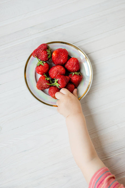 Hand of a child with a strawberry on a rustic background, a plate of strawberries. - Фото, изображение