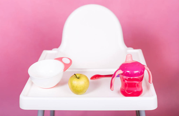 White baby chair for eating with an apple spoon and a cup on a bright colored light pink background - Foto, Bild