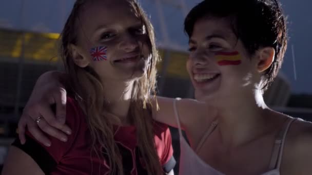 Dwie młode dziewczyny, fani piłki nożnej w deszczu, Anglii i Hiszpanii, uśmiechając się, śmiejąc się, obejmując, stadion w tle 50 fps - Materiał filmowy, wideo