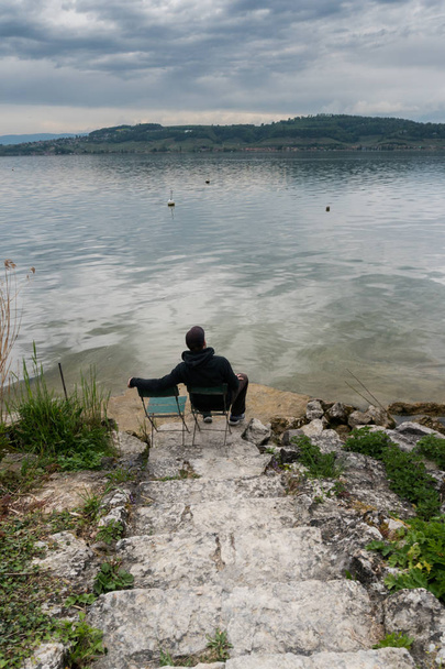 muž sedí na židli u krásného jezera na klidné jarní večer s skalní schody vedoucí dolů mu - Fotografie, Obrázek