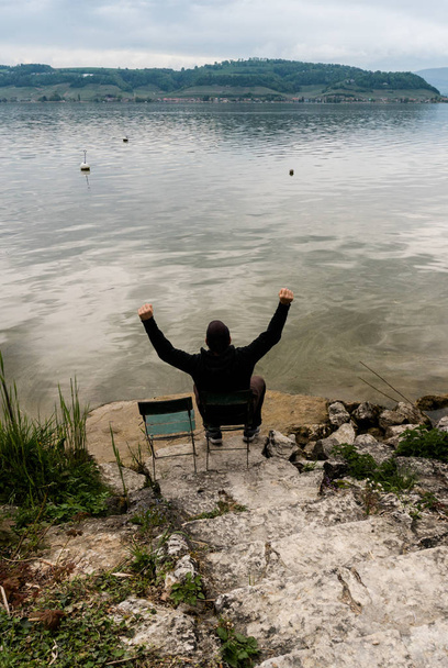 hombre sentado en una silla en las orillas de un hermoso lago en una tranquila primavera, con los brazos levantados en celebración y alegría
 - Foto, Imagen