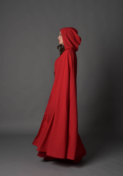 Ganztägiges Porträt einer brünetten Dame in rotem Fantasiekostüm mit Umhang, stehend posierend auf grauem Studiohintergrund. - Foto, Bild