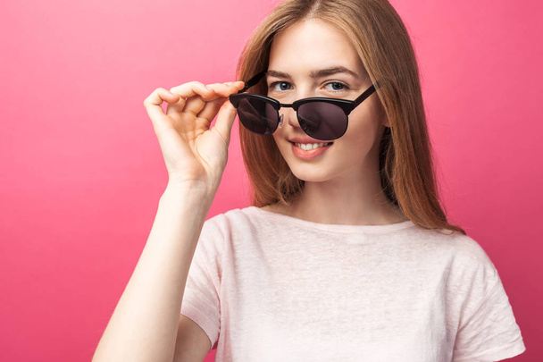Beautiful young blonde girl in glasses standing on pink background wearing jeans, pink top smiling snow white smile, wearing black glasses and looking perfect promotional photo - Photo, image