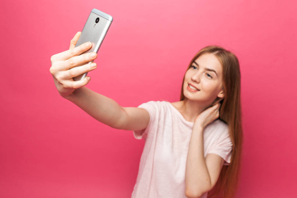 Portrait of cheerful beautiful girl, photographed, taking selfie, looking into the phone, funny enough, isolated on pink background, advertising - Valokuva, kuva