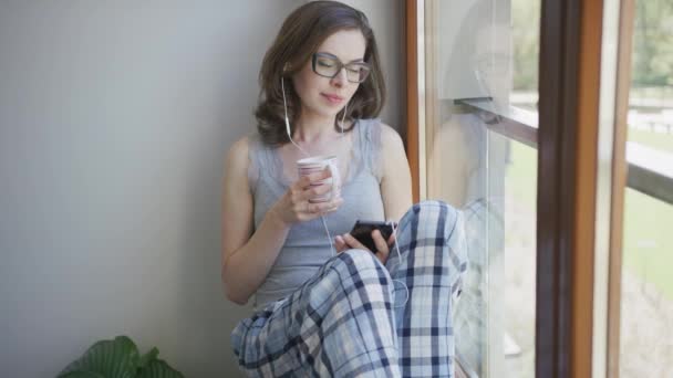 Mujer joven sentada por la ventana usando un teléfono inteligente con auriculares
 - Metraje, vídeo