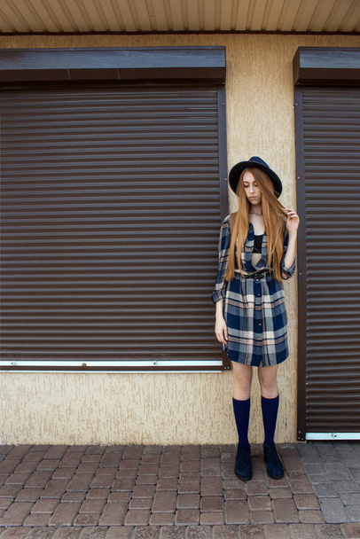 Red hair girl in plaid shirt and vintage hat, with brown backpack pose on the street. Fashion and stylish concept. - Photo, Image