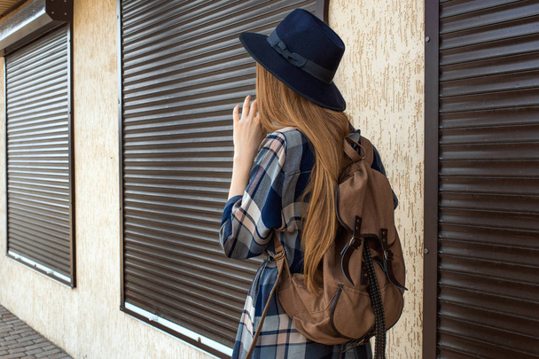 Chica de pelo rojo en camisa a cuadros y sombrero vintage, con pose mochila marrón en la calle. Moda y concepto elegante
. - Foto, imagen
