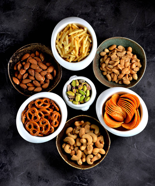 Different kinds of snacks - chips, salted peanuts, cashews, almonds and pistachios, pretzels with salt, potatoes, salted straw. - Photo, Image