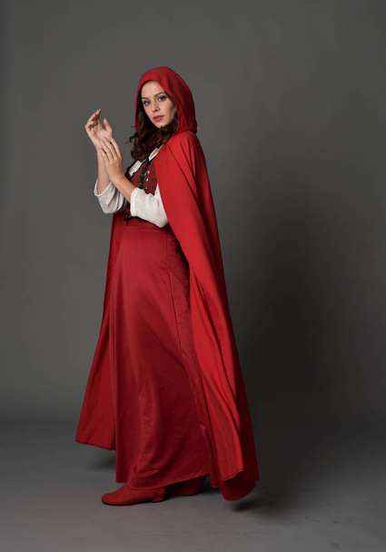 Ολόσωμο πορτρέτο της μελαχρινή κοπέλα φορώντας κόκκινο φαντασίας κοστούμι με μανδύα, στέκεται πόζα σε φόντο γκρι στούντιο. - Φωτογραφία, εικόνα