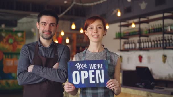Pretty red-haired businesswoman cafe owner is holding "yes we are open" sign with her employee in apron standing near her. Successful start-up and people concept. - Footage, Video