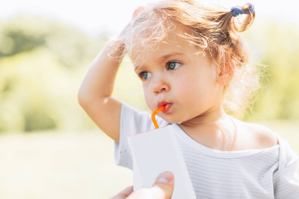 Sunny portrait of a little child drinking from a straw juiceon blurred summer background - Фото, изображение
