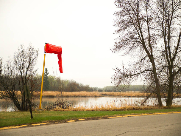 Κάλτσα άνεμος φωτεινό κόκκινο ύφασμα στο πάνω μέρος του ένα κίτρινο καλάμι κρέμεται limp κατά μήκος της ακτογραμμής του ποταμού σε ένα ήρεμο πρωινό σε Μινεσότα Warroad με δέντρα και συννεφιά λευκή και γκρι πέρα από. - Φωτογραφία, εικόνα