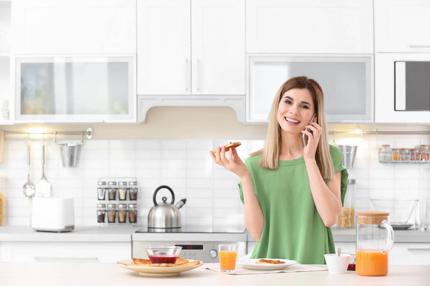 Beautiful woman eating tasty toasted bread with jam while talking on mobile phone at table in kitchen - Photo, Image