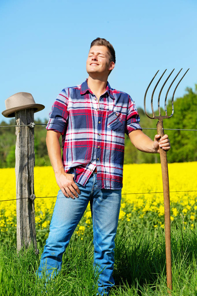 handsome blond man with pitchfork in front of yellow field  - Zdjęcie, obraz