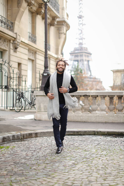 Afro american male person running with Eiffel Tower in background, wearing grey scarf and black sweater. - Φωτογραφία, εικόνα