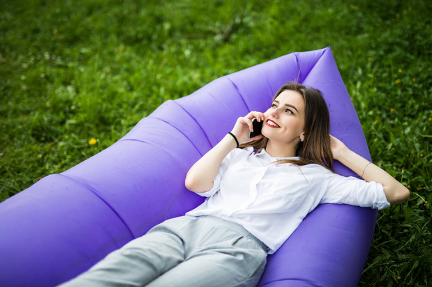 Красивая молодая женщина лежит на надувном диване lamzac говорить по телефону во время отдыха на траве в парке
 - Фото, изображение