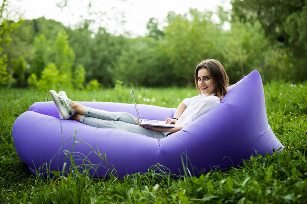 Pretty young woman lying on inflatable sofa lamzac browse in internet on laptop while resting on grass in park - Photo, image