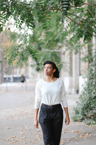 Afro american kobieta ładne spacery w mieście, w pobliżu zielonych drzew i budynku, na sobie białą bluzkę. - Zdjęcie, obraz