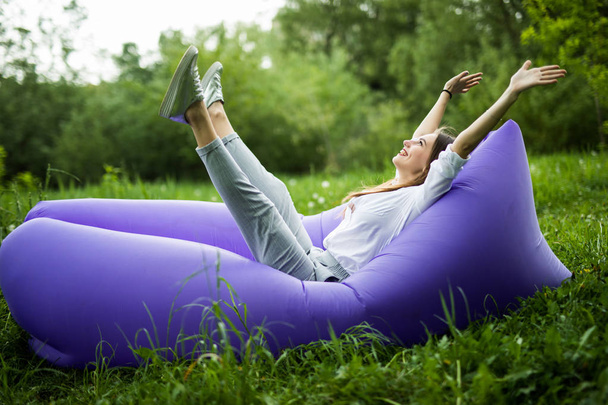 I am free. Pretty young woman lying on inflatable sofa lamzac with reised hands in the air while resting on grass in park - 写真・画像