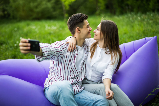 あなたは私の魂です。若いカップルがハグ、インフレータブルソファ lamzac と selfie 公園の芝生の上で休憩の携帯電話上に座ってキス - 写真・画像