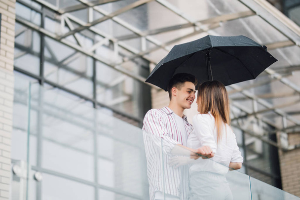 Young couple of lovers under a umbrella in city. Handsome people having fun in city urban center in a raining day. Love and vacation concept - Photo, Image