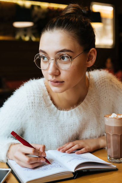 pensive young girl freelancer with glasses sitting in a cafe, working, writing in her notebook - Foto, Imagen