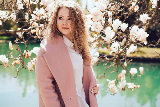 curly-haired woman wearing a pink coat posing next to a blooming fragrant magnolia in a park, ranks with a pond - Zdjęcie, obraz