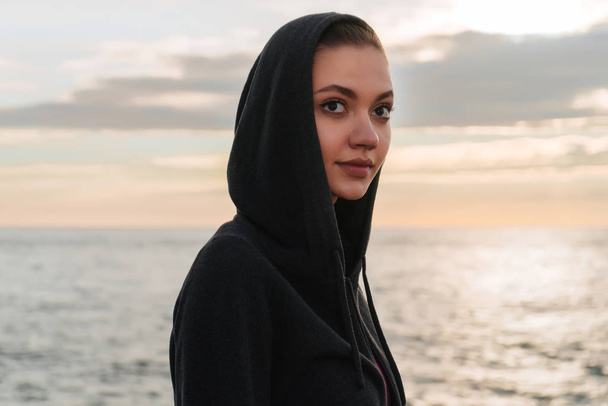 young beautiful girl in a hood resting after jogging by the sea, at sunset - Fotografie, Obrázek