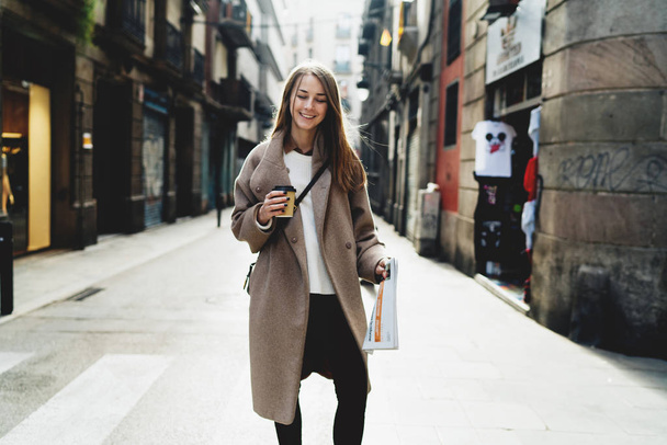 Cheerful young woman with take away coffee and newspaper walking on the street  - Foto, Imagem