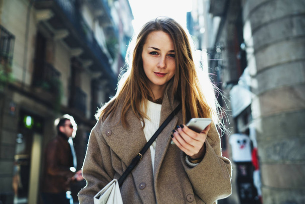 Half length portrait of young woman holding a mobile phone in her hand looking at the camera on blurred city street background - Photo, image