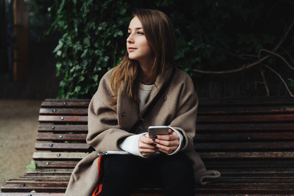 Young woman in a trendy outfit using smartphone while sitting on wooden bench outdoors - Foto, afbeelding