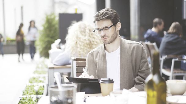 A handsome european man in eyeglasses making notes at cafe outdoors - Foto, imagen