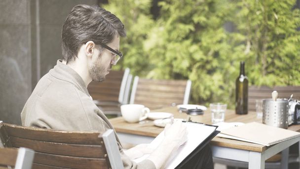 A handsome european man in eyeglasses making notes drawing at cafe outdoors - Фото, изображение