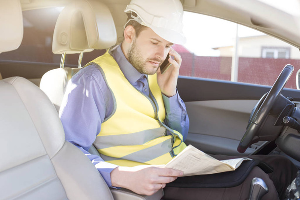 Male construction engineer in white helmet, shirt and yellow waistcoat sits in the car and scans documents while talking on the phone - Φωτογραφία, εικόνα