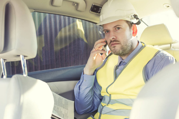 Male construction engineer in white helmet, shirt and yellow waistcoat sits in the car and scans documents while talking on the phone - Photo, Image