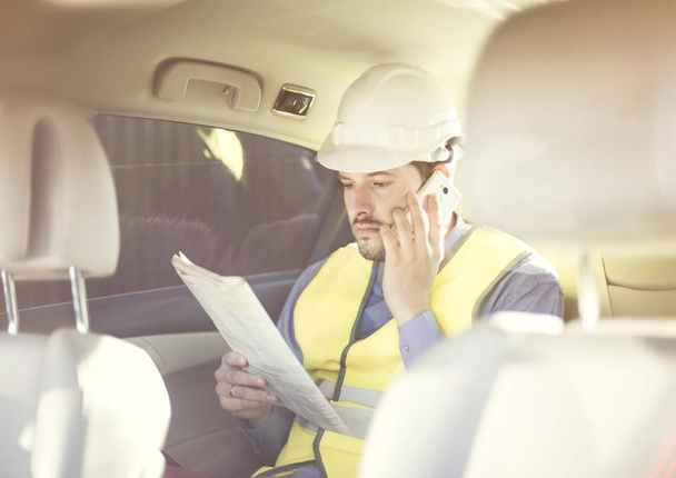Male construction engineer in white helmet, shirt and yellow waistcoat sits in the car and scans documents while talking on the phone - Photo, Image