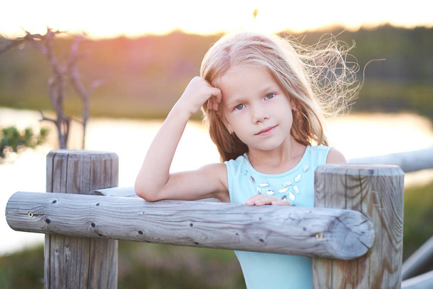 Portrait of a cute little girl, standing outdoors while leaning on a wooden fence. - Foto, Bild