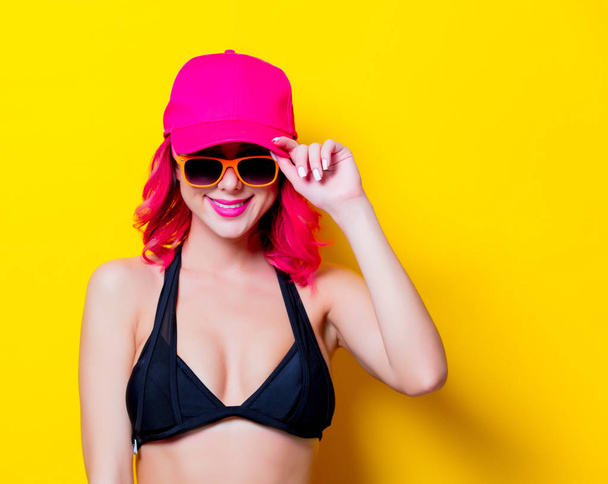 Joven chica de pelo rosa en bikini y gafas naranjas con gorra. Retrato aislado sobre fondo amarillo
 - Foto, imagen