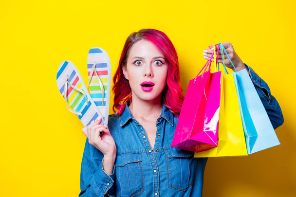 Young pink hair girl in blue shirt holding a colored shopping bags and flip flops. Portrait on isolated yellow background - Photo, Image
