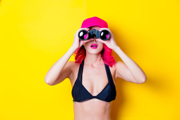 junges rosafarbenes Mädchen im Bikini mit Mütze und Fernglas. Porträt isoliert auf gelbem Hintergrund - Foto, Bild