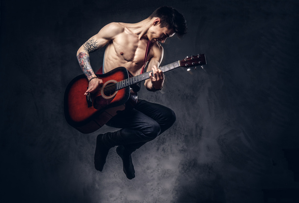 ジャンプしながらギターを弾くハンサムな上半身裸の若い音楽家. - 写真・画像