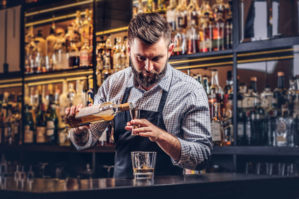 Κομψό βάναυση μπάρμαν σε ένα πουκάμισο και ποδιά κάνει ένα κοκτέιλ στο μπαρ μετρητή φόντο. - Φωτογραφία, εικόνα